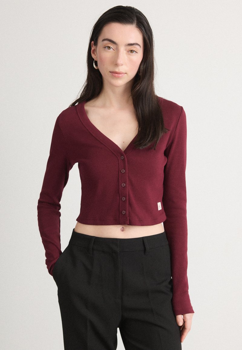 Cardigan scurt de culoare burgundă, cu mâneci lungi, textură ribben și detalii cu butoni pe față. Asortat cu pantaloni negri, cu talie înaltă.