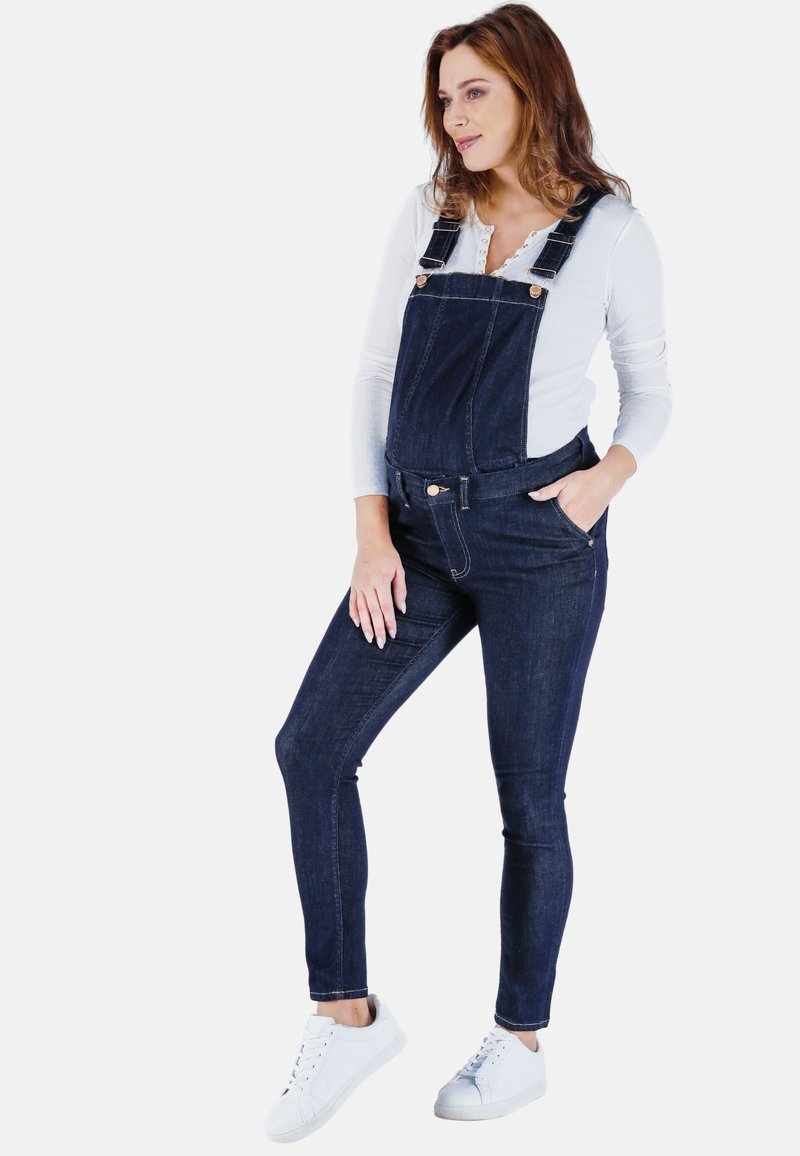 Donkerblauwe denim overall met verstelbare schouderbanden, voorkennis en aansluitende pijpen, gecombineerd met een witte lange mouwtop en witte sneakers.