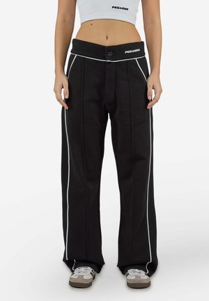 Femme portant un pantalon noir à jambes larges avec passepoil blanc et taille boutonnée, associé à un débardeur court blanc et des baskets.