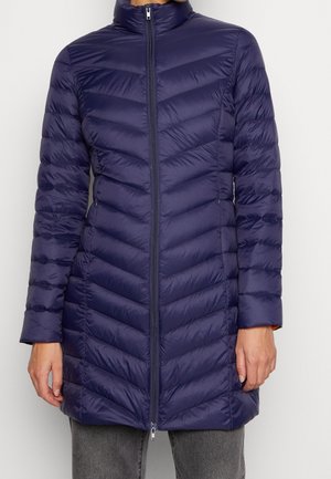 Marineblauwe gewatteerde pufferjas met chevronstiksels, volledige frontrits, hoge kraag en lange mouwen, gedragen over grijze broek.