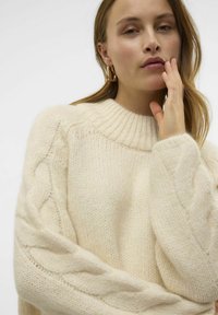 Cremefarbener, gestrickter Pullover mit geripptem Ausschnitt und Kabelstrickdetails an den Ärmeln. Weiche Textur und oversized Passform.