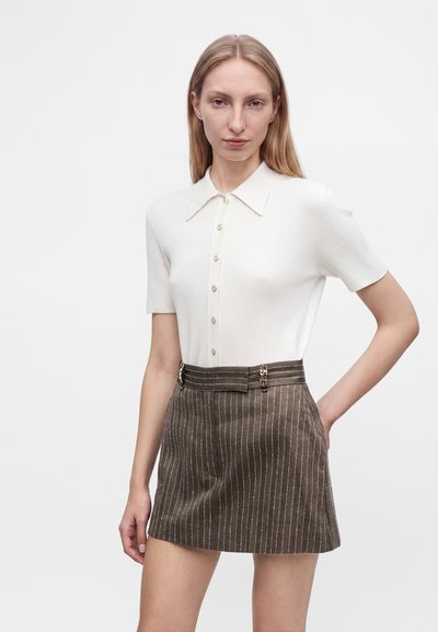 Femme aux cheveux blonds raides porte un haut blanc boutonné à manches courtes et une jupe marron à fines rayures en ligne A avec des détails de fermoir.