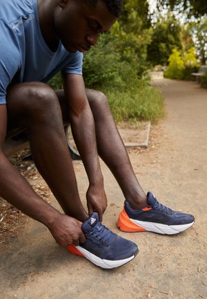 Jeune homme assis sur un banc en plein air, attachant ses chaussures de course bleu marine et orange sur un chemin de terre entouré de verdure.