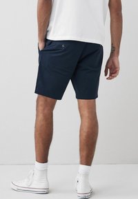 Pantalones cortos azul marino de un material ligero, con un diseño de frente plano, bolsillos laterales y un bolsillo trasero con cierre de botón. Combinados con zapatillas blancas.