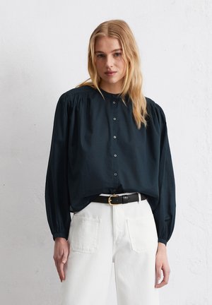 Donkergroene blouse met ballonsmouwen, zacht materiaal, ronde halslijn, gecombineerd met witte high-waisted broek en een zwarte riem.