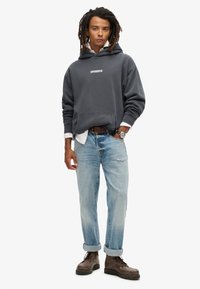 Sudadera gris con capucha y logo blanco, llevada sobre una camisa blanca, combinada con jeans azul claro arremangados en los puños y botas marrones.