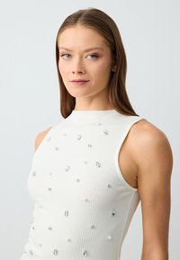 Top bianco a coste senza maniche con collo alto, caratterizzato da decorazioni sparse in cristallo e simili a perle per un tocco di texture e dettaglio.