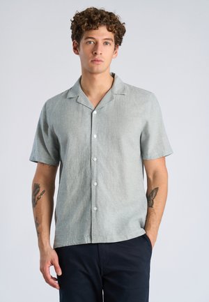 Młody mężczyzna o kręconych włosach, ubrany w jasnoszarą koszulę na krótki rękaw z guzikami i ciemne spodnie, stojący z jedną ręką w kieszeni.