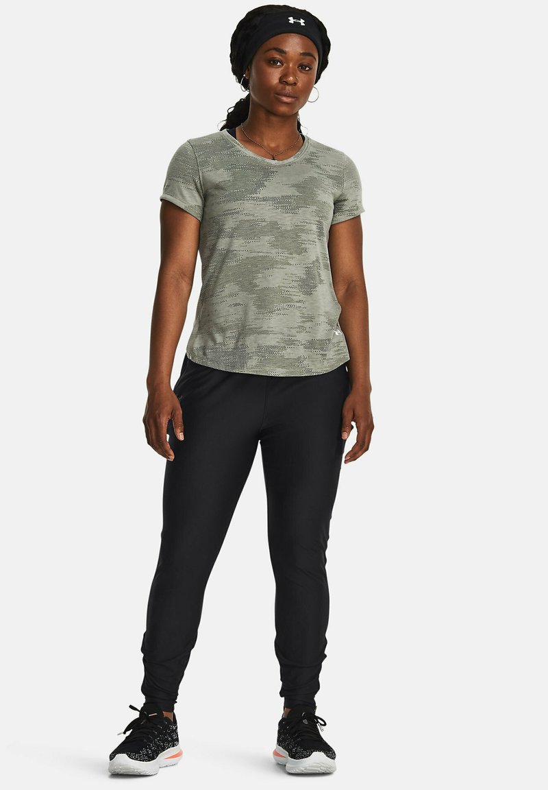 Maglietta sportiva verde mimetica a maniche corte, abbinata a pantaloni neri attillati e sneakers nere con dettagli bianchi.