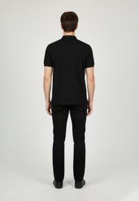 Czarna koszulka polo z krótkim rękawem i kołnierzykiem, w połączeniu z czarnymi spodniami. Gładka faktura i dopasowany krój. Widok z tyłu pokazuje ogólny zarys sylwetki.