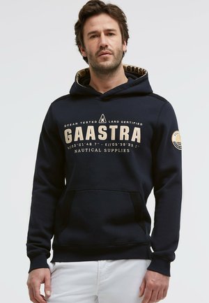 Mann trägt einen marineblauen Gaastra-Kapuzenpullover mit dem Schriftzug "Nautical Supplies" und Koordinaten, kombiniert mit weißen Hosen, steht vor einem schlichten Hintergrund.