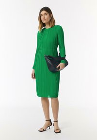 Robe plissée verte à manches longues et col rond, assortie à un sac à main noir en courbe et des talons à lanières noires. Texture lisse.