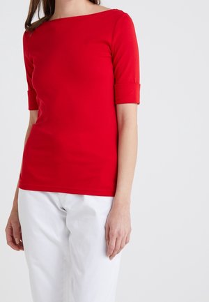 Kobieta ubrana w dopasowaną czerwoną koszulę z rękawami do łokcia i białe spodnie, stojąca na jednolitym jasnym tle.