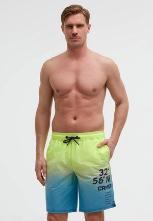 Hemdloser Mann mit kurzen Haaren, der gelb-blaue Farbverlauf-Badeshorts mit dunklem blauen Kordelzug und bedrucktem Text am linken Oberschenkel trägt.