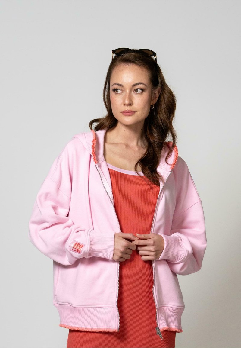 Felpa rosa chiaro con zip, dotata di polsini e orlo a costine, caratterizzata da un bordo sfrangiato. Indossata sopra a un vestito arancione brillante, mette in evidenza una vestibilità rilassata.