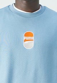 Sudadera azul claro con un logotipo bordado de PUMA en naranja y blanco en el centro. Presenta un cuello redondo de canalé y una textura suave.