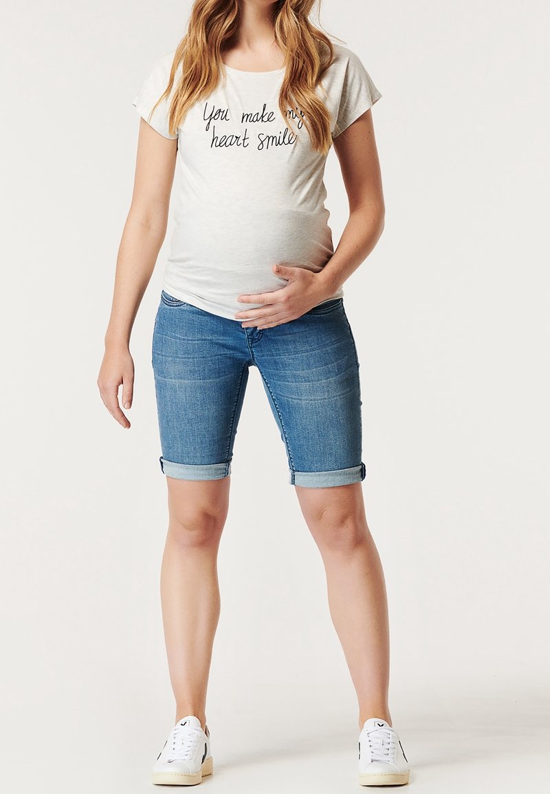 Femme enceinte portant un t-shirt blanc avec texte et un short en jean bleu retroussé, tenant son ventre d'une main.