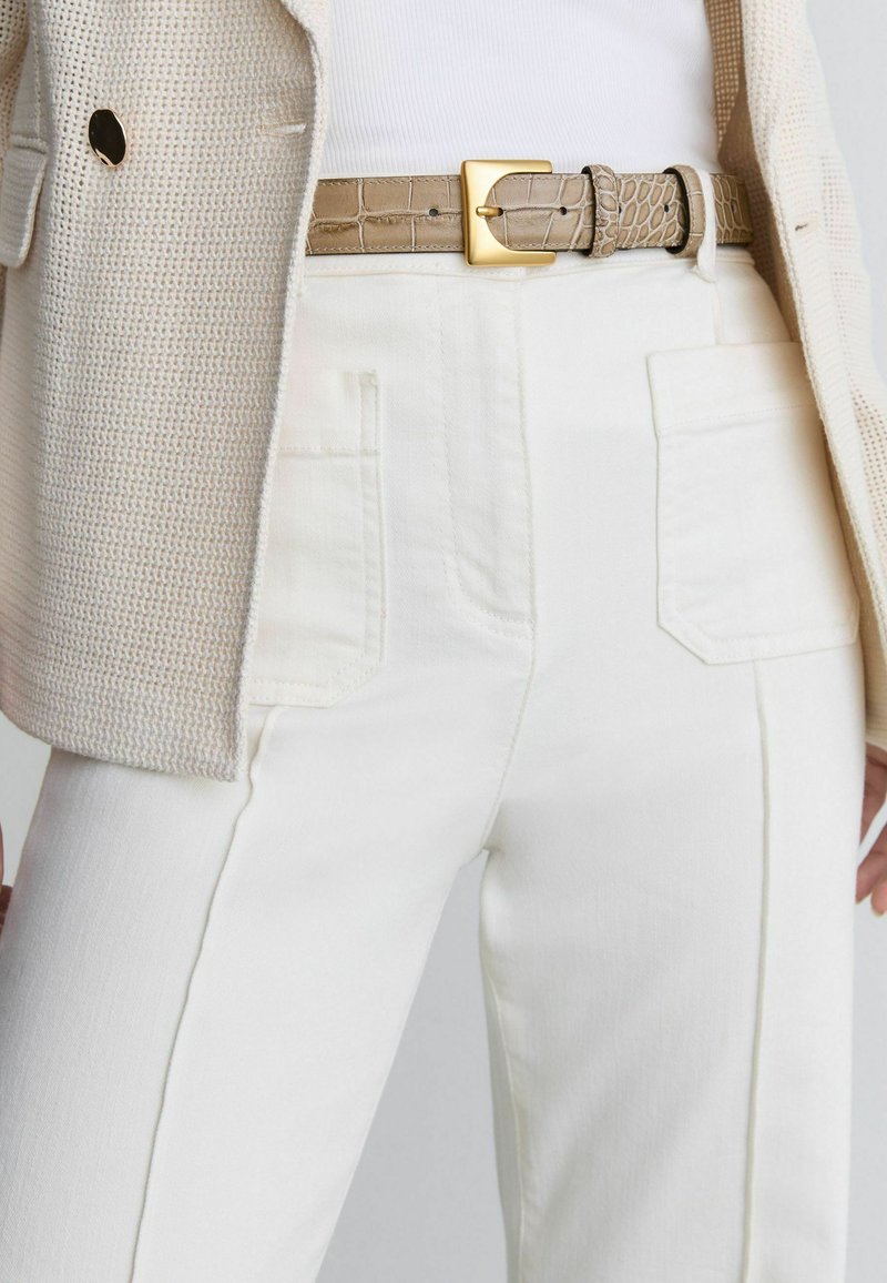 Primer plano de blazer con textura crema, pantalones blancos de talle alto con bolsillos delanteros y cinturón beige con patrón de cocodrilo y hebilla dorada.