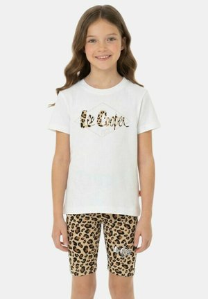 Fille souriante aux longs cheveux bruns portant un t-shirt blanc Lee Cooper et un short à imprimé léopard, debout devant un fond uni.