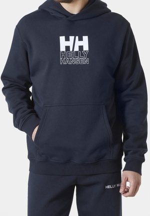 Mężczyzna ubrany w granatową bluzę z kapturem i spodnie marki Helly Hansen, bluza posiada przednią kieszeń i duże białe logo na piersi.
