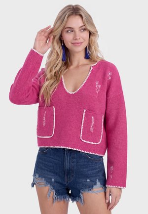Kvinde med langt blond hår iført en pink V-hals sweater med hvidt blomsterbroderi og revnede denimshorts, poserer med hånden nær hovedet.