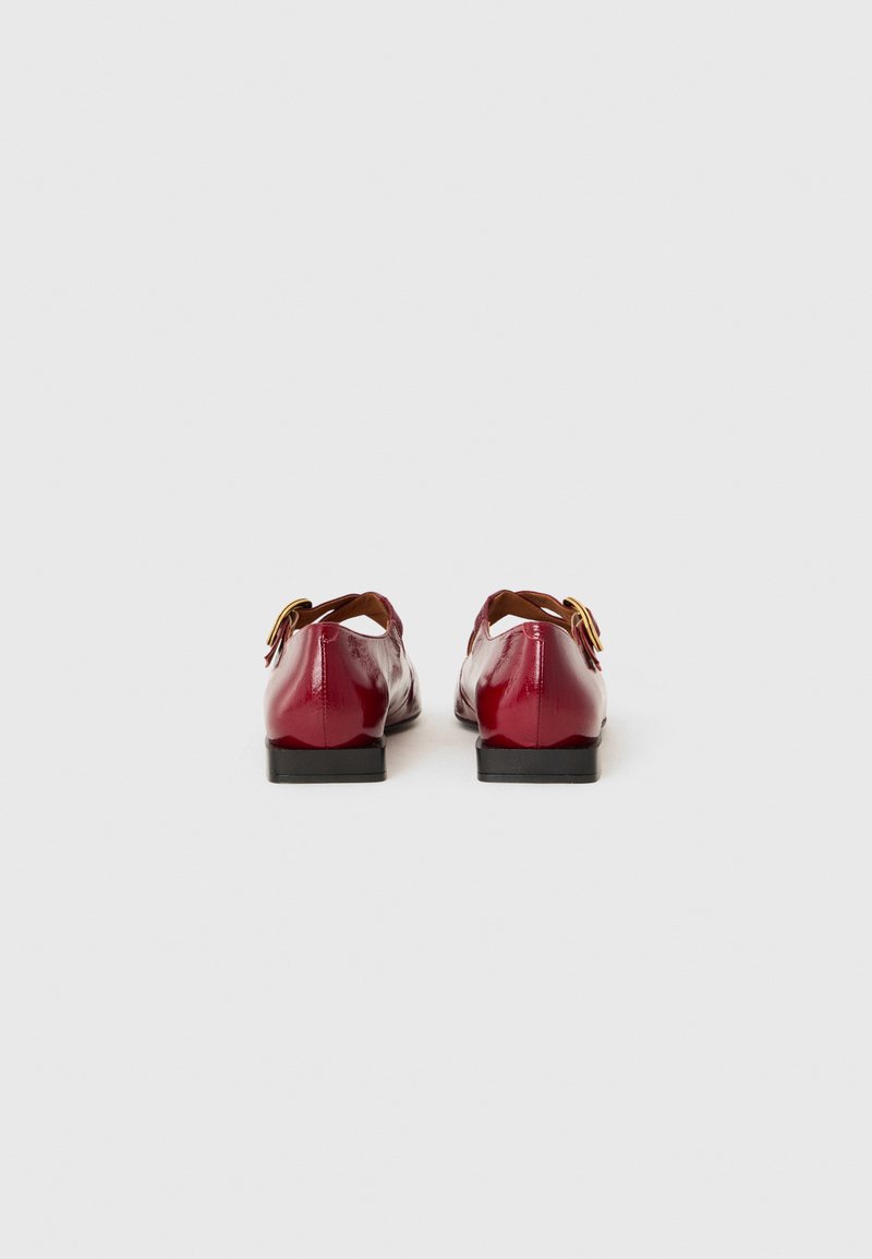 Scarpe in vernice rossa con tacco basso nero e una cintura regolabile beige con fibbia effetto oro, caratterizzate da un design elegante e arrotondato.