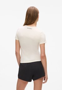 Tricou cu mânecă scurtă de culoare albă-crem, cu un design ajustat, având un logo discret pe spate. Materialul pare neted și elastic. Asortat cu pantaloni scurți negri.
