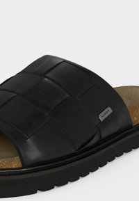 Μαύρο δερμάτινο σανδάλι slide με πλεκτό πάνω μέρος, φελλό πάτο, παχιά ελαστική σόλα και μικρή μεταλλική ετικέτα μάρκας στο πλάι.