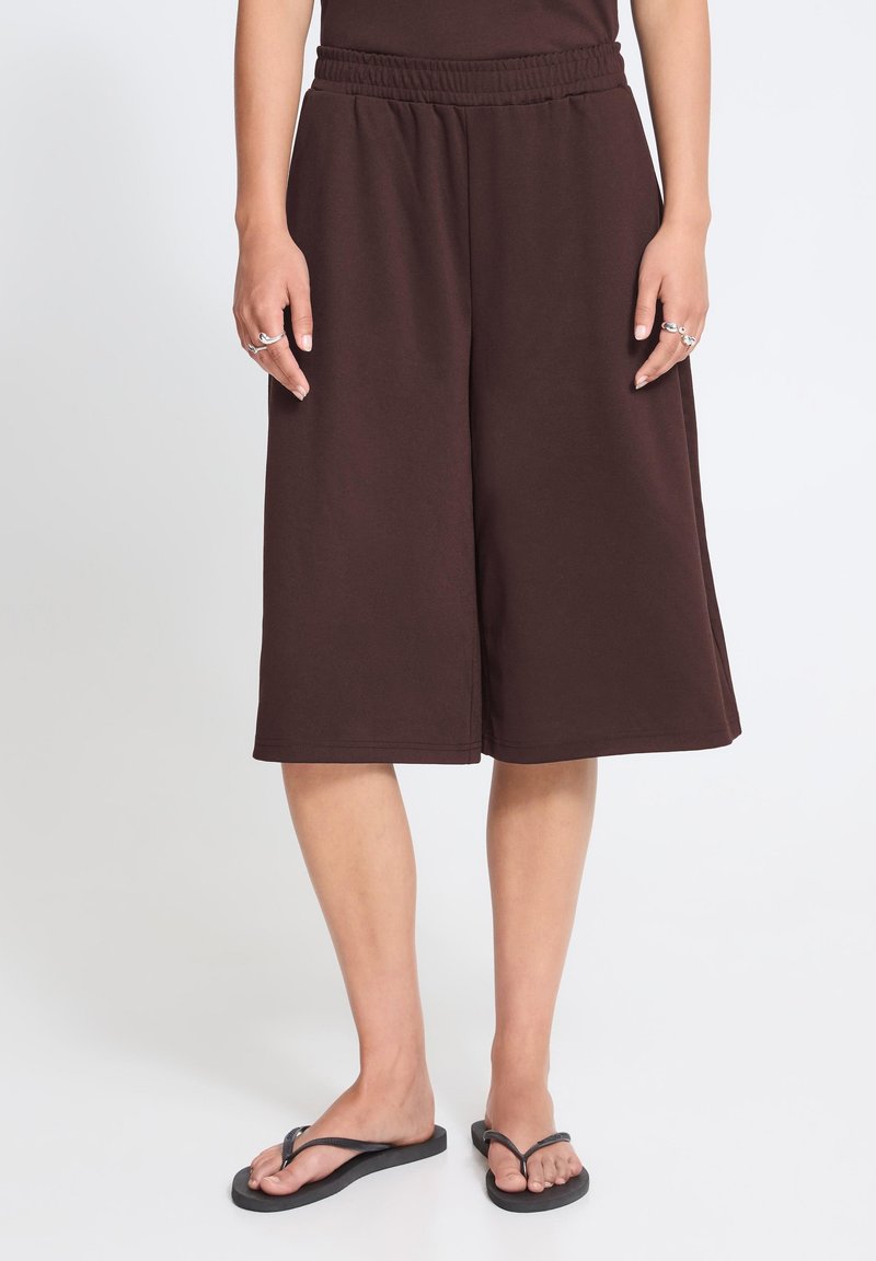Personne portant un short ample marron foncé jusqu'aux genoux, des tongs noires et des bagues aux deux mains sur un fond uni clair.