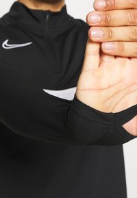 Black athletic top with a high collar and zip closure, featuring a prominent white Nike logo on the chest and thumbholes in the sleeves.