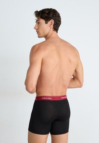 Zwarte katoenen boxer briefs met een bordeauxrood elastische tailleband met het Calvin Klein-logo, weergegeven vanaf de achterkant op een model.