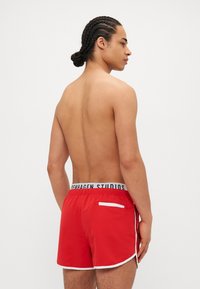Red swim shorts with a white trim and a back pocket, featuring a black elastic waistband with "Copenhagen Studios" text.