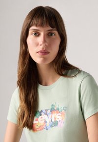 Lys grønn bomull t-skjorte med blomstermønster og teksten "Levi's" i hvitt. Rund hals og korte ermer, glatt tekstur.