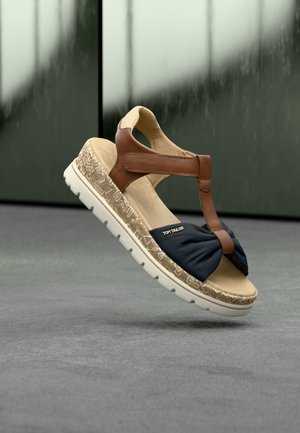 Sandale bleu marine avec un dessus torsadé, des brides en cuir brun et une semelle blanche texturée avec des motifs géométriques. Logo de la marque visible.