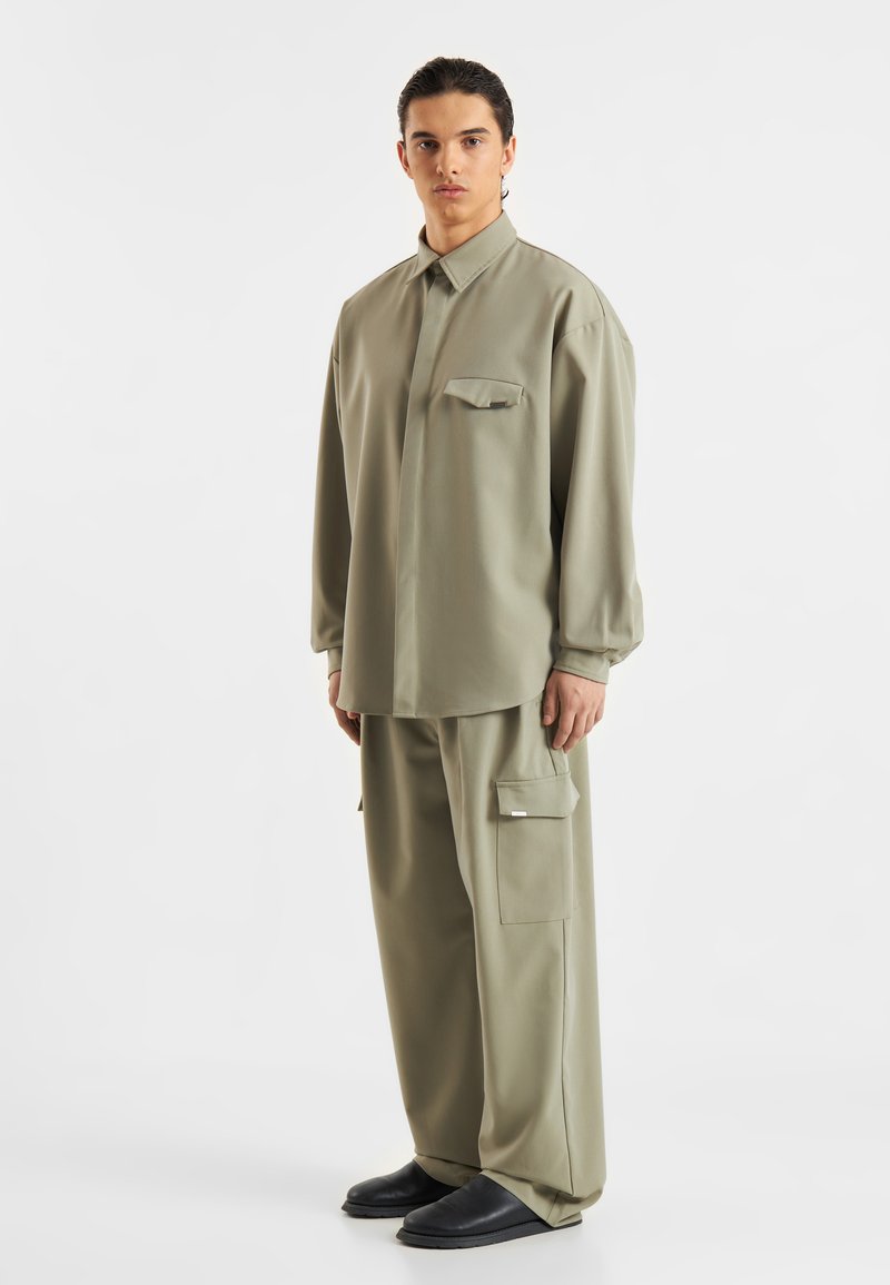 Khaki srajca z gumbi in enim žepom na prsih, v kombinaciji s širokimi hlačami z bočnimi žepi in gladko, mat teksturo.