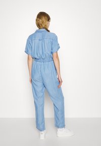 Kvinna som står med ryggen mot kameran, klädd i en ljusblå denimoverall och vita sneakers mot en enkel vit bakgrund.