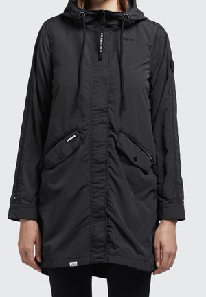 Kobieta ubrana w czarną, lekką kurtkę z kapturem, wyposażoną w skośne kieszenie na zamek i na zatrzaski, z zamkiem błyskawicznym z przodu i sznurkami do ściągania.