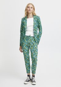 Blommig kostym i grönt, blått och gult, med en figursydd kavaj och avsmalnande byxor. Matchas med vit t-shirt och svarta sneakers.