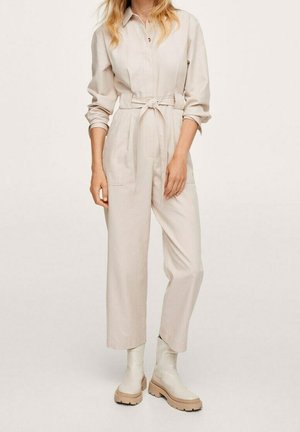 Beżowy kombinezon z kołnierzykiem zapinanym na guziki, długimi rękawami, paskiem w talii i dwiema przednimi kieszeniami. Noszony z kremowymi botkami do kostek na grubych podeszwach.