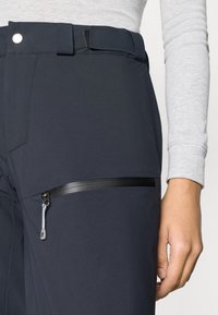 Μαύρο παντελόνι softshell με φερμουάρ σε τσέπη, θηλιές ζώνης και λεία υφή, με ένα χέρι να ακουμπά στην περιοχή της μέσης.