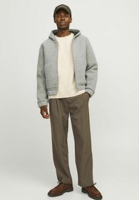 Grauer Reißverschluss-Hoodie über einem beigen Pullover, kombiniert mit braunen Hosen und braunen Schuhen. Der Hoodie hat eine weiche Textur und einen lockeren Schnitt.