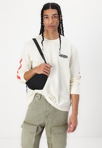 Sort crossover taske, beige skjorte med lange ærmer og rød grafik på ærmerne, grøn cargo-bukser med store lommer, båret afslappet.