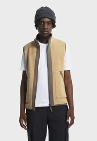 Beige Fleece-Weste mit braunen Akzenten, Reißverschluss vorne und zwei seitlichen Taschen, getragen über einem weißen T-Shirt, vor einem neutralen Hintergrund.