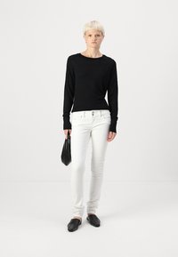 Svart langermet genser med naglede detaljer, hvite slim-fit jeans og svarte slip-on sko. Modellen holder en liten svart håndveske.