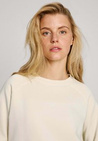 Sudadera de color crema con cuello redondo y mangas raglán. Presenta un sutil logo en la parte frontal y una textura suave y lisa.