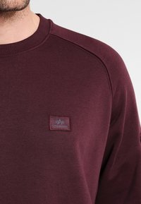 Bordeauxrotes Sweatshirt mit Rundhalsausschnitt aus weichem Stoff; verfügt über einen kleinen grauen Logoaufnäher auf der Brust sowie gerippte Bündchen und Saum.