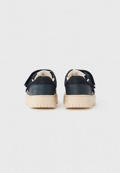 Barnens marinblå sneakers med beige sulor och kardborreband, sedd bakifrån mot en enkel vit bakgrund.