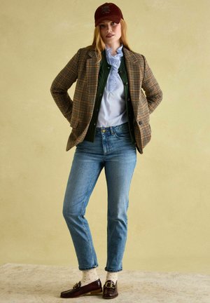 Brown plaid blazer over a green cardigan, light blue shirt, and blue jeans. Maroon cap with logo, brown loafers, and polka-dotted socks.