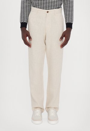 Personne portant un pantalon en lin beige, des baskets blanches et un haut à manches longues à motif noir et blanc, debout devant un fond blanc uni.