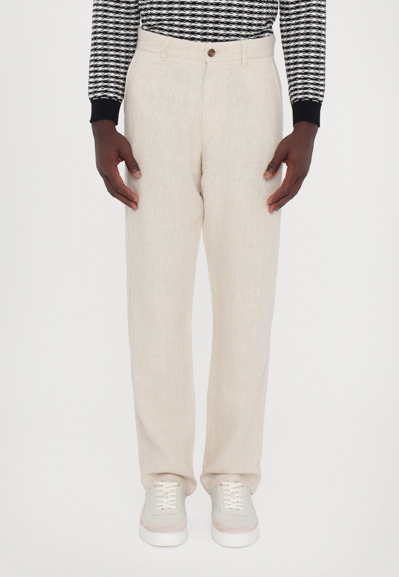 Person trägt beige Leinenhose, weiße Sneakers und ein schwarz-weiß gemustertes langärmeliges Oberteil, steht vor weißem Hintergrund.